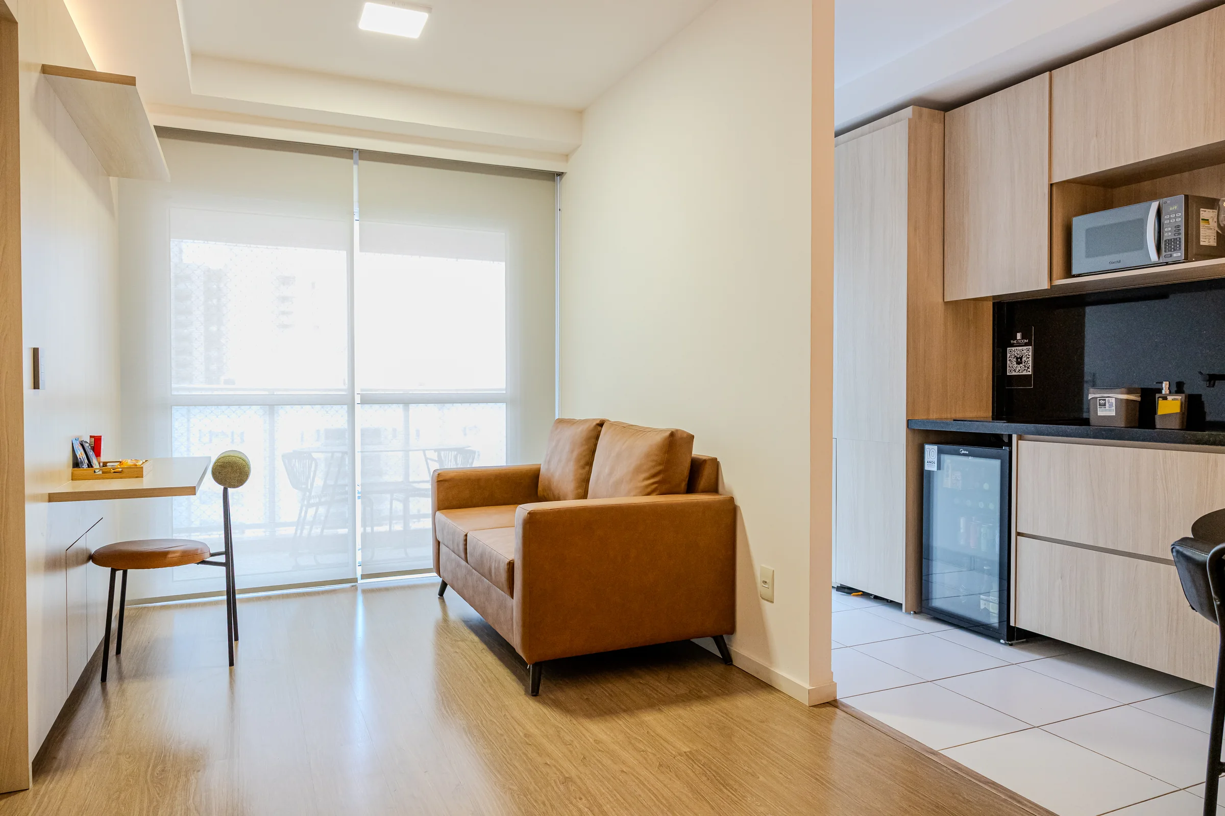 Sala iluminada com sofá, bancada junto à janela e cozinha ao lado.