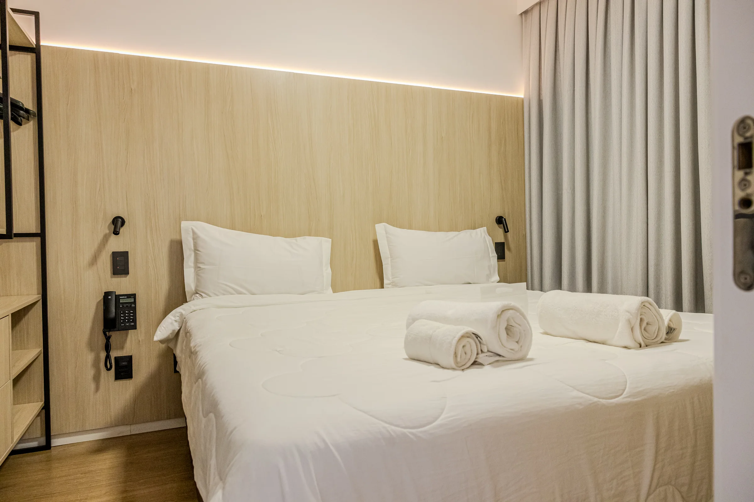 Quarto com cama de casal, toalhas brancas e cortina cinza.