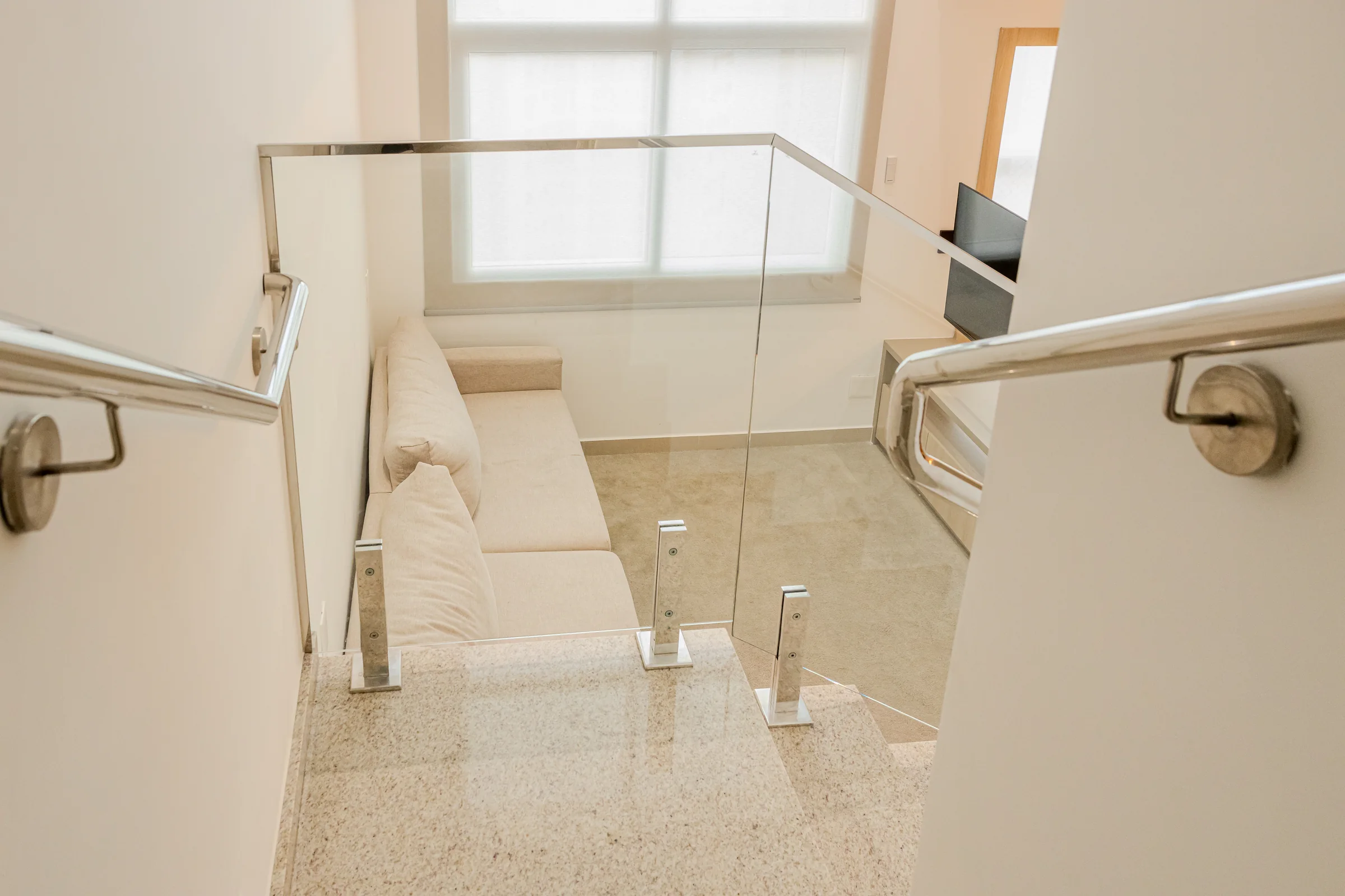 Sofá e sala vistos do alto da escada com guarda-corpo de vidro.