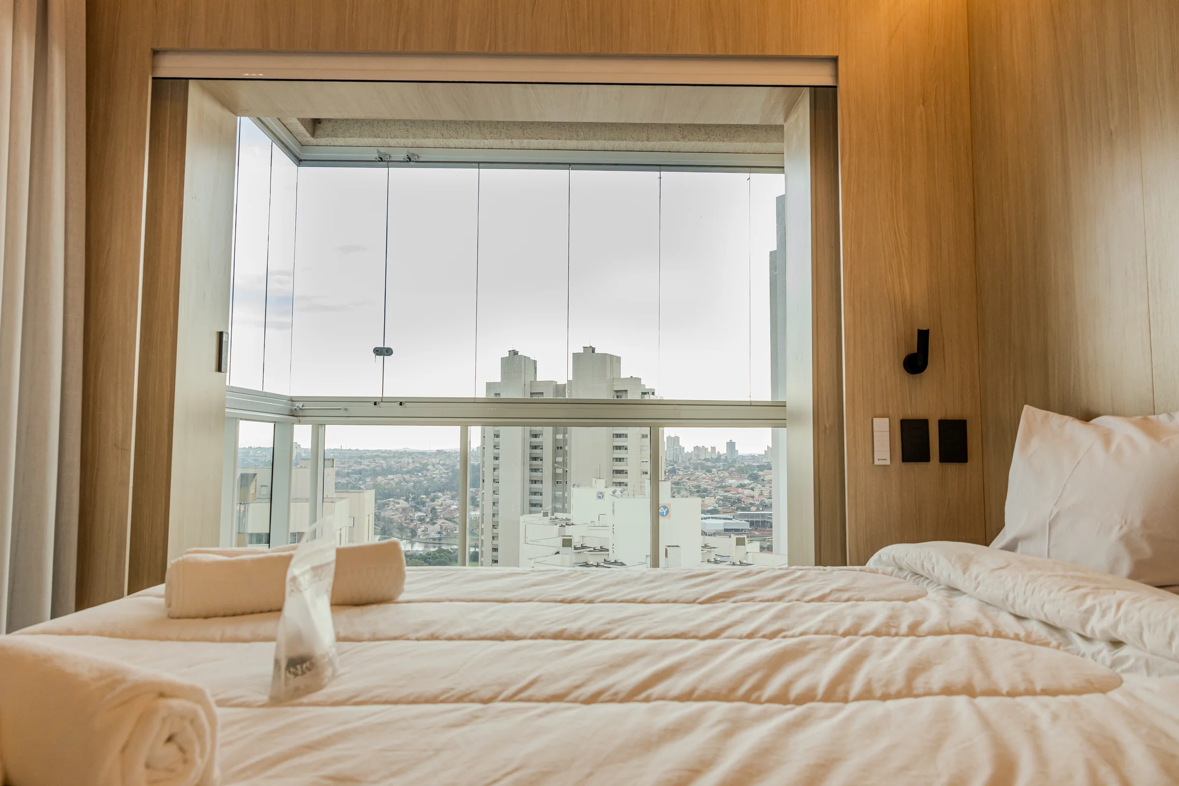 Cama de casal diante de uma grande janela com vista urbana.