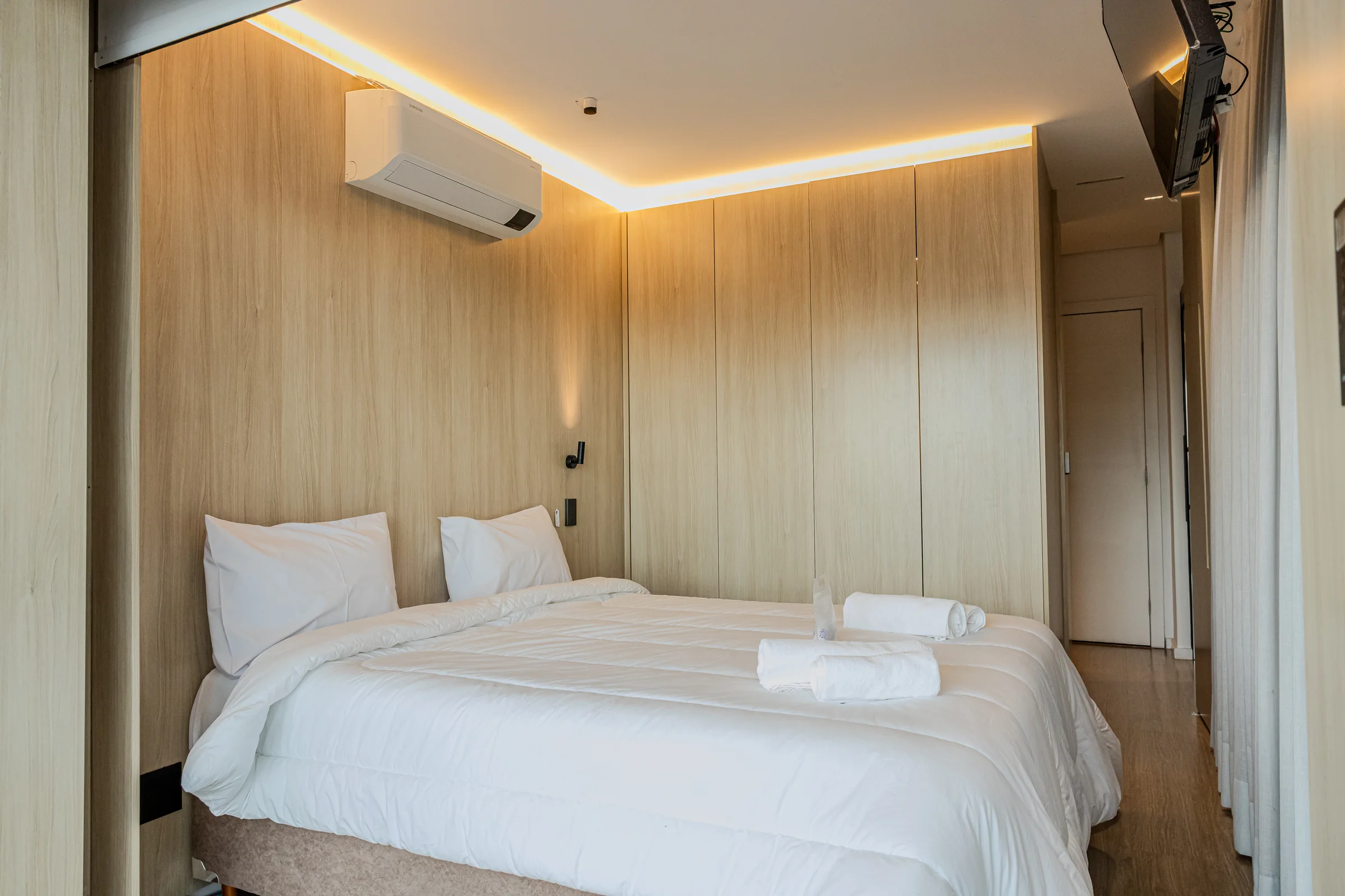 Quarto principal com cama, iluminação indireta e passagem lateral.